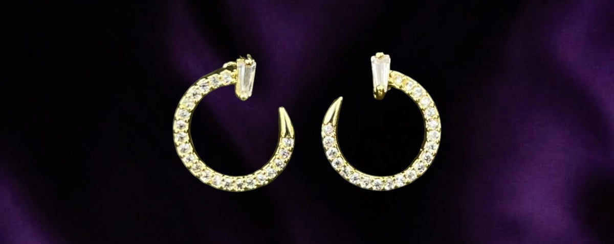 SHE BEAUTY Crescent Luxe gold-tone hoop earrings with baguette and round crystal accents on a purple silk background