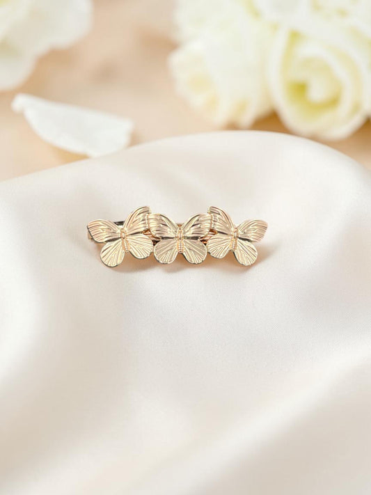 SHE BEAUTY metallic gold butterfly barrette hair clip with intricate wing detailing on a soft cream fabric background.