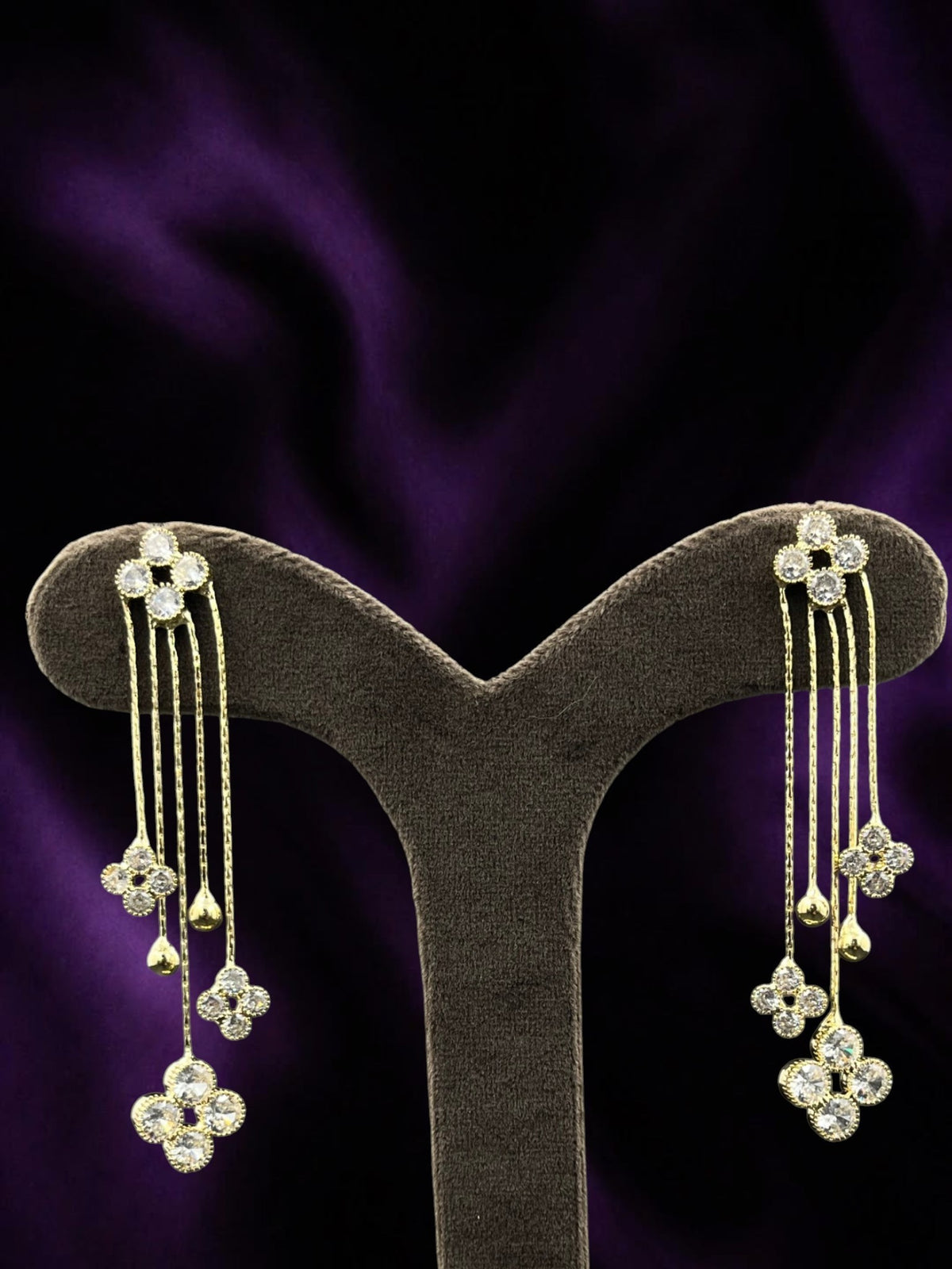 SHE BEAUTY Floral Trails gold-tone drop earrings with sparkling clear stone flower accents on a dark purple background