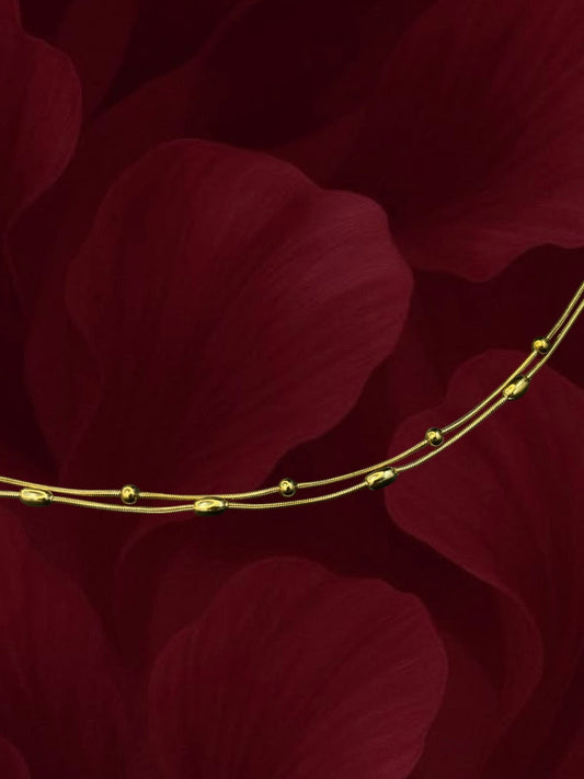 SHE BEAUTY KIYU ZIYU gold-toned double-strand anklet with delicate beads against a dark red floral background.