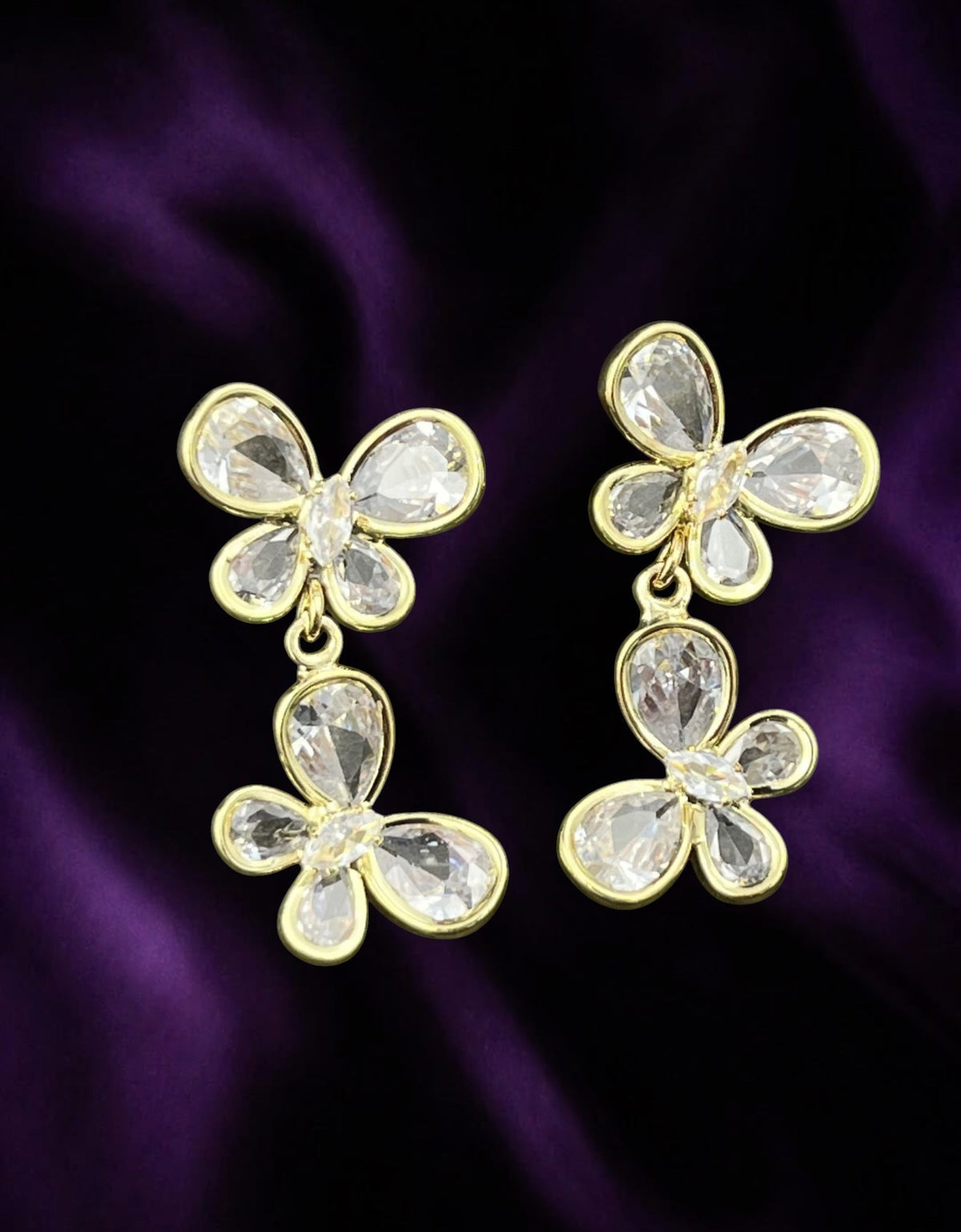 SHE BEAUTY gold-tone crystal butterfly drop earrings featuring sparkling clear stones on a dark purple satin background