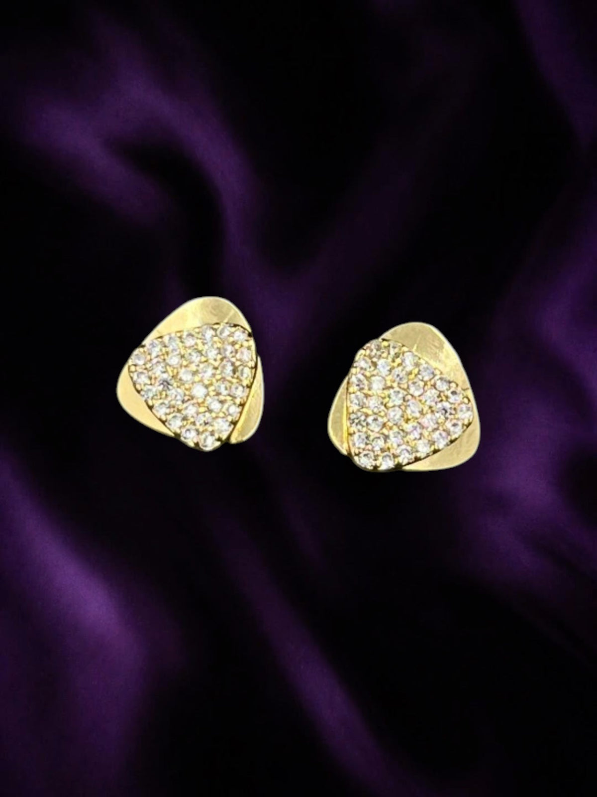 SHE BEAUTY gold-toned triangle stud earrings with sparkling pave crystals on a deep purple satin background