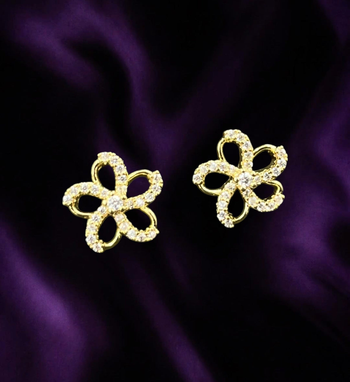 SHE BEAUTY gold-toned flower stud earrings with sparkling cubic zirconia stones on a deep purple satin background