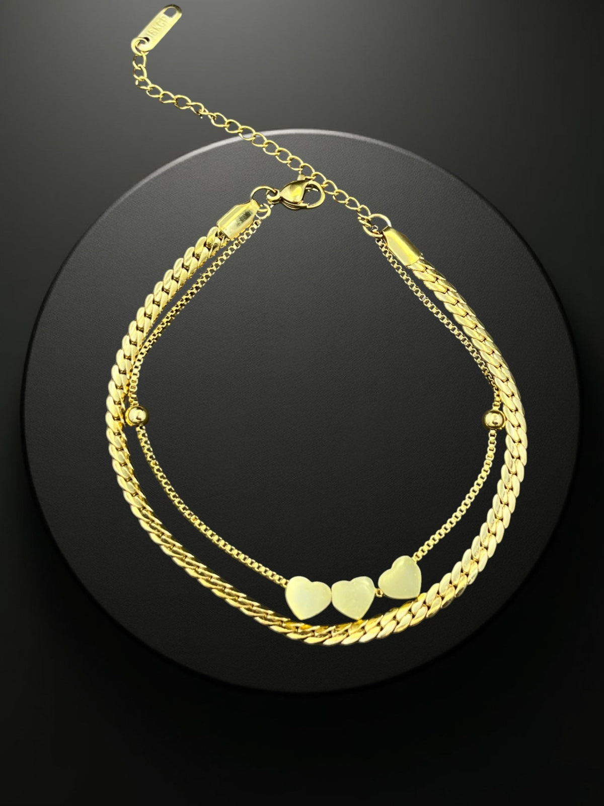 SHE BEAUTY KIYU ZIYU gold-toned double-layer chain bracelet featuring three heart charms on a black display stand.