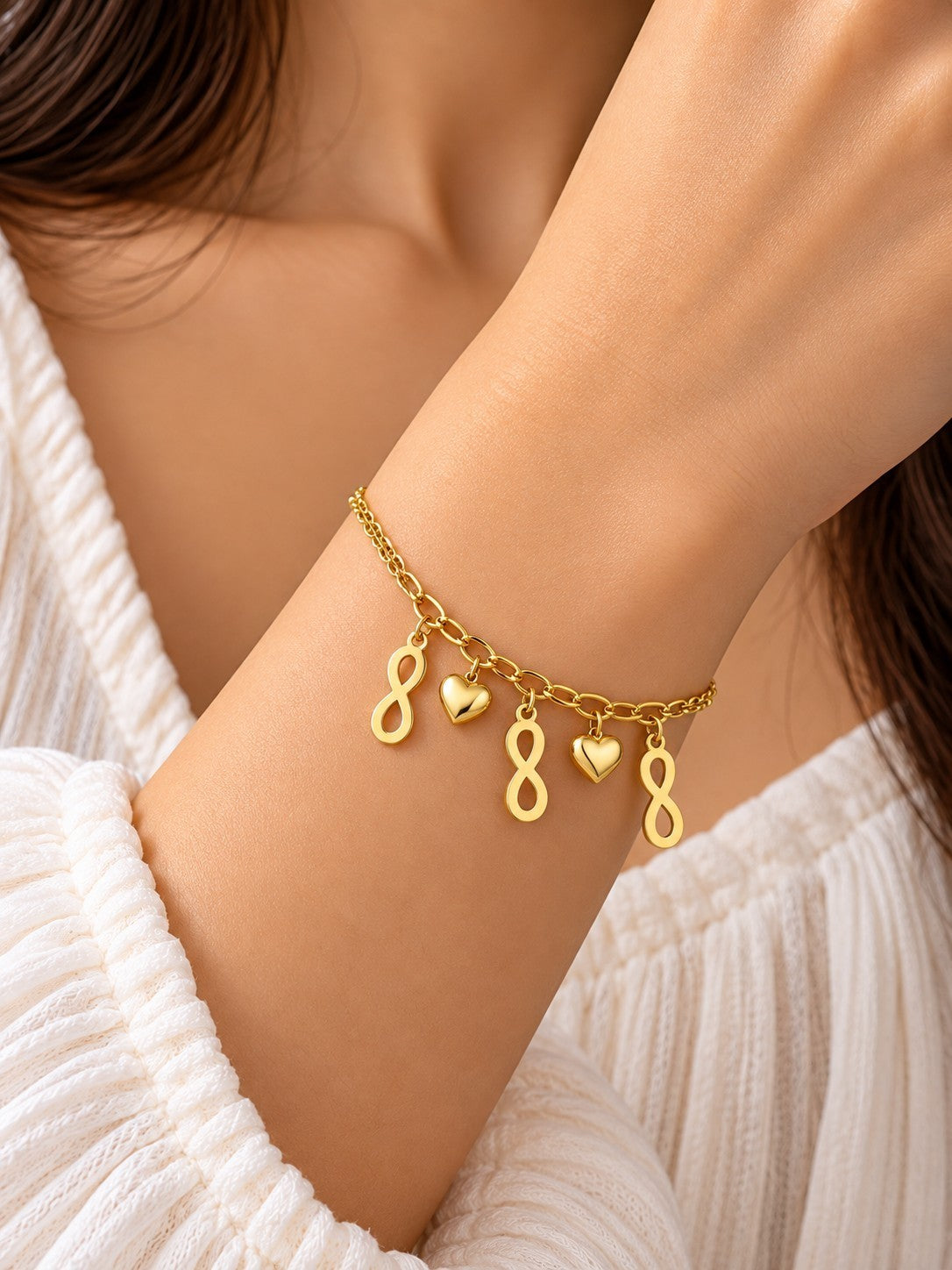 SHE BEAUTY gold-tone chain bracelet featuring infinity symbol and heart charms on a woman's wrist