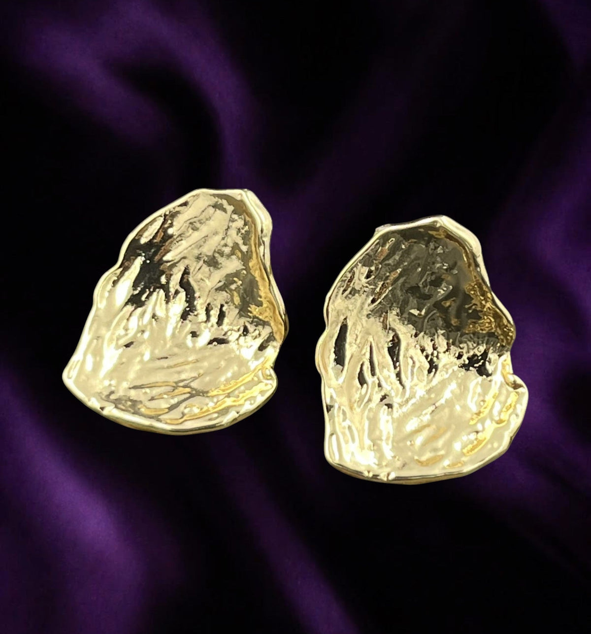 SHE BEAUTY anti-tarnish gold-toned earrings with a textured, organic abstract shape on a deep purple satin background.