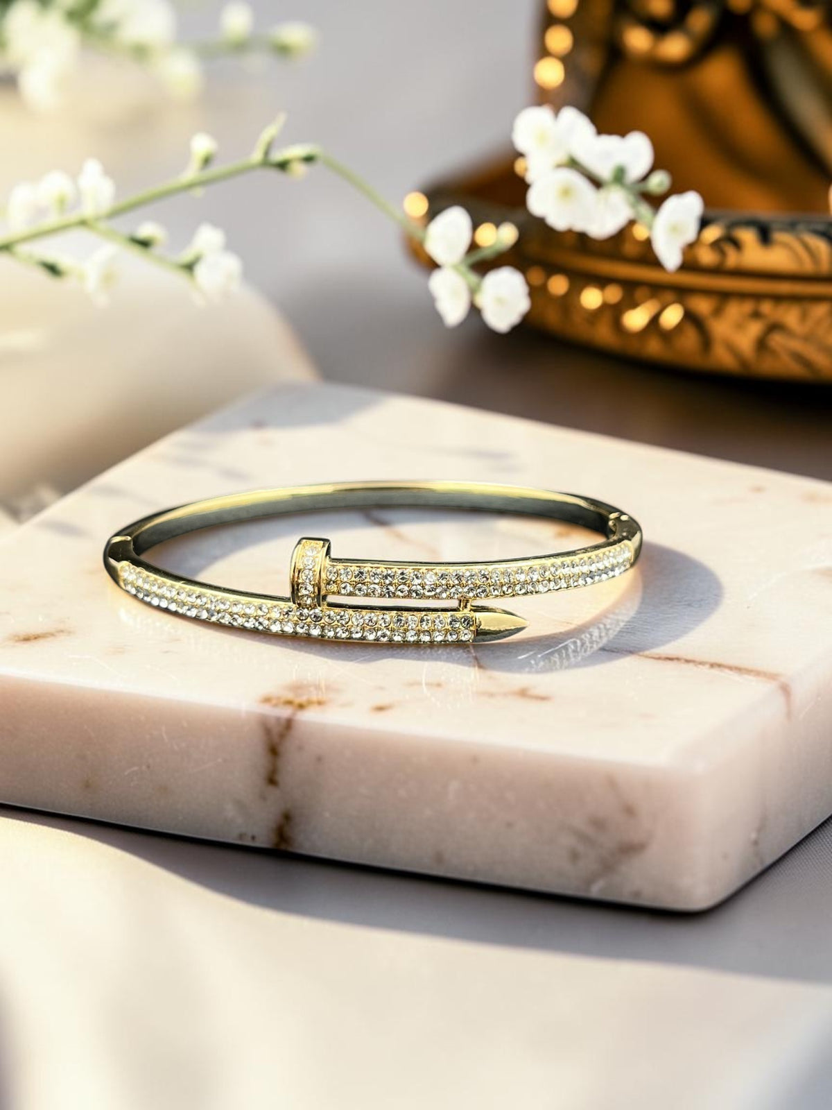 SHE BEAUTY gold-toned nail-style bangle bracelet encrusted with sparkling zirconia stones on a marble display block.