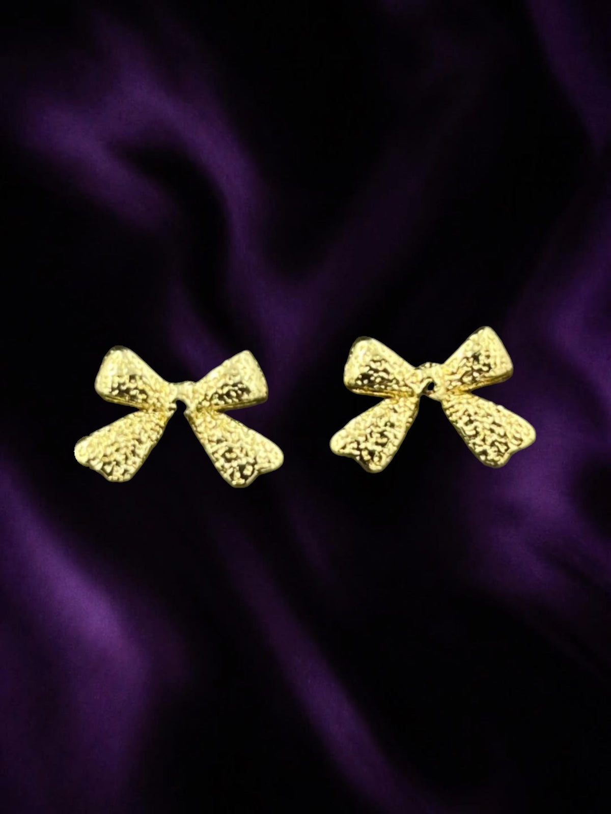 SHE BEAUTY GOLDEN BOW earrings featuring a textured gold finish on a dark purple satin background