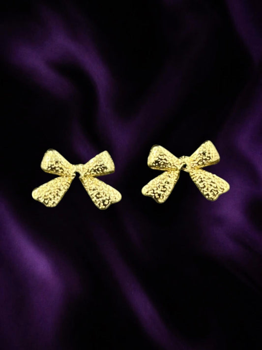 SHE BEAUTY GOLDEN BOW earrings featuring a textured gold finish on a dark purple satin background