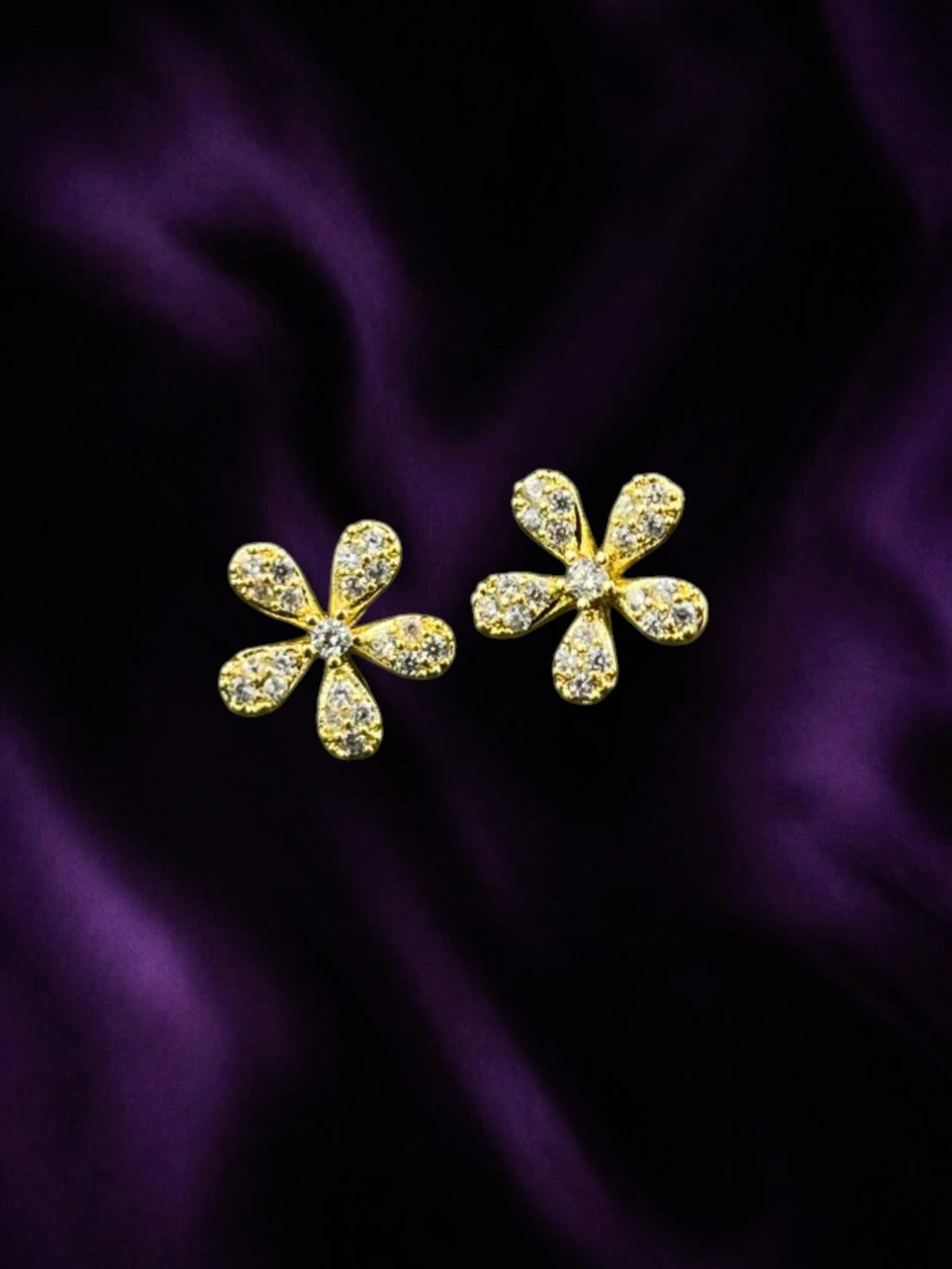 SHE BEAUTY LUNA PATELS gold-toned flower stud earrings with sparkling white stones on a dark purple satin background