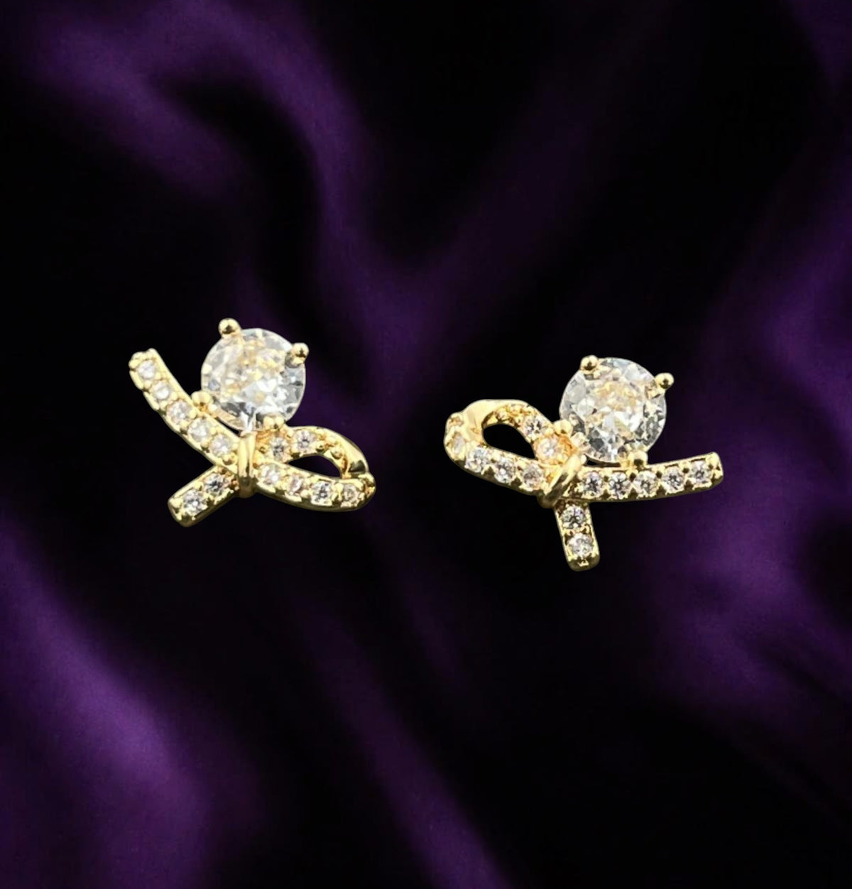 SHE BEAUTY LUXE KNOT STUDS featuring gold-tone ribbon bows with sparkling clear crystals on a purple satin background
