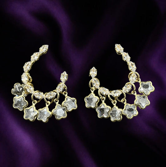SHE BEAUTY STARRY LOOPS gold-tone crescent earrings with dangling star-shaped crystals on a purple satin background